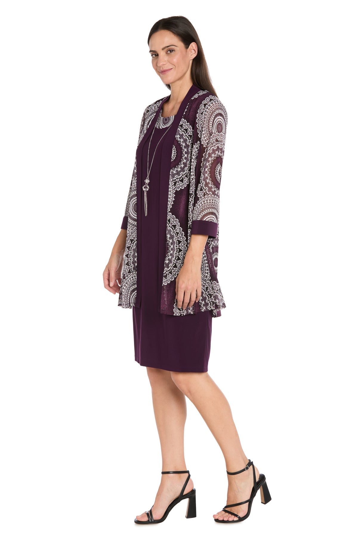 A woman wears the R&M Richards 9626P Petite Two-Piece Short Party Dress, featuring a matching jacket, black heels, and a long necklace, as she stands sideways against a white background.
